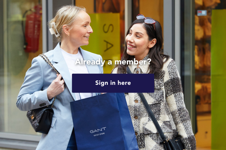 Two women smile at each other in Helsinki Outlet Village. One is carrying a blue GANT Outlet Store shopping bag. Text over the image reads: "Already a member? Sign in here".