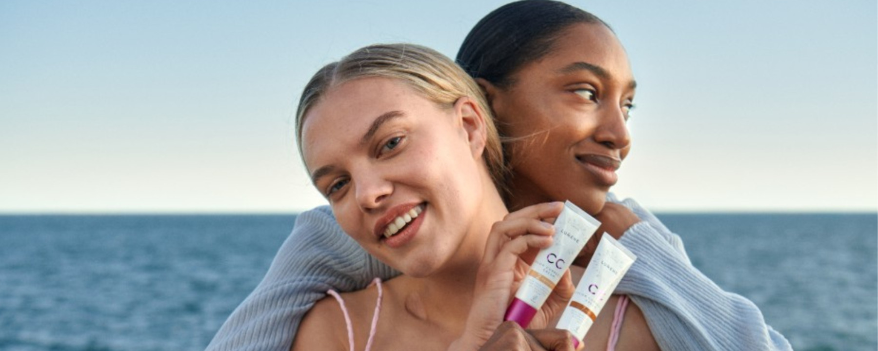 Two women are hugging each other and showing off tubes of foundation. The sea is in the background.