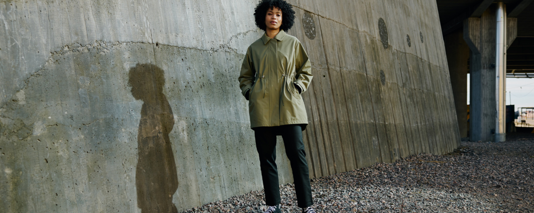 A woman stands on a sandy gravel road wearing a green parka