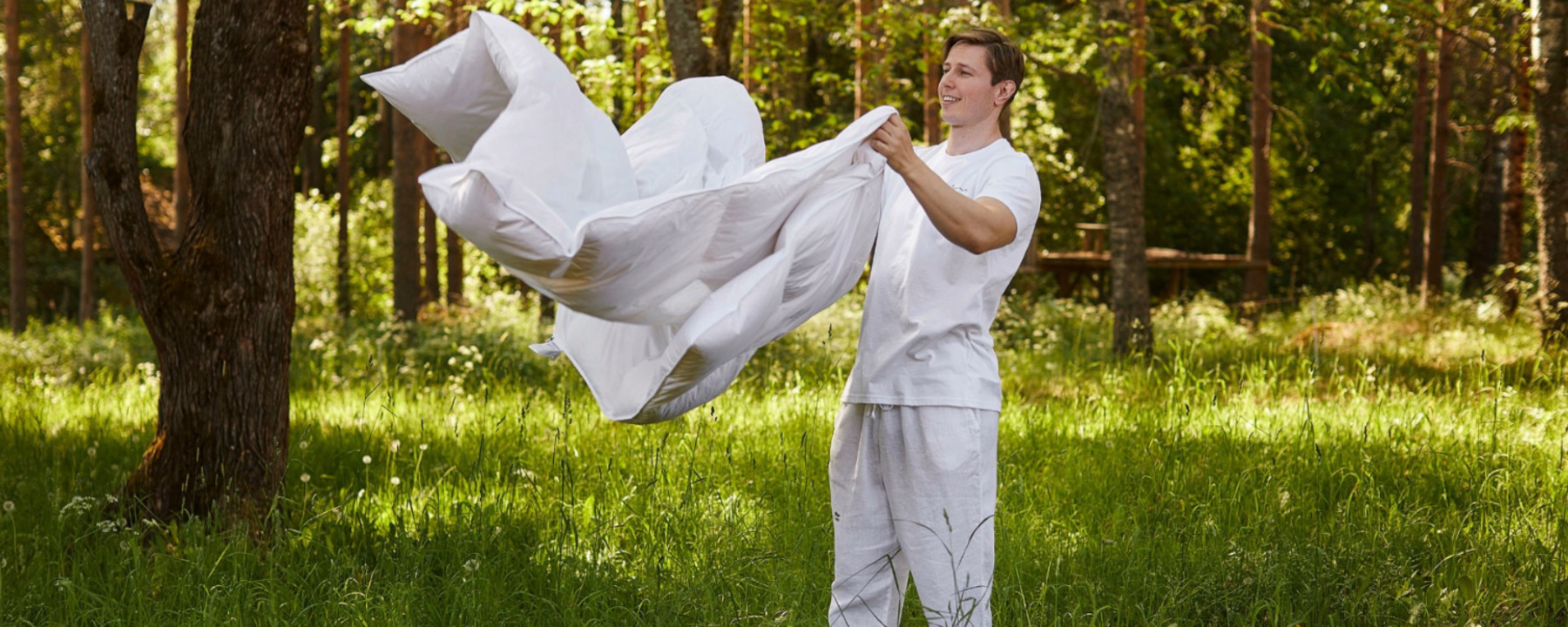 A man wearing white clothes raises a white blanket into the air in the forest.