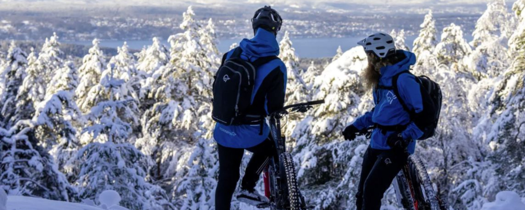 A man and a woman on bicycles on top of a snowy fell, wearing blue Norrøna jackets