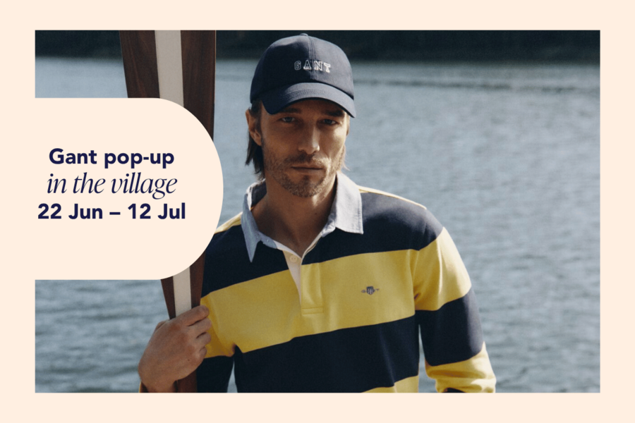 A man wearing a dark blue GANT cap and a striped GANT shirt is holding an oar and standing by the water.