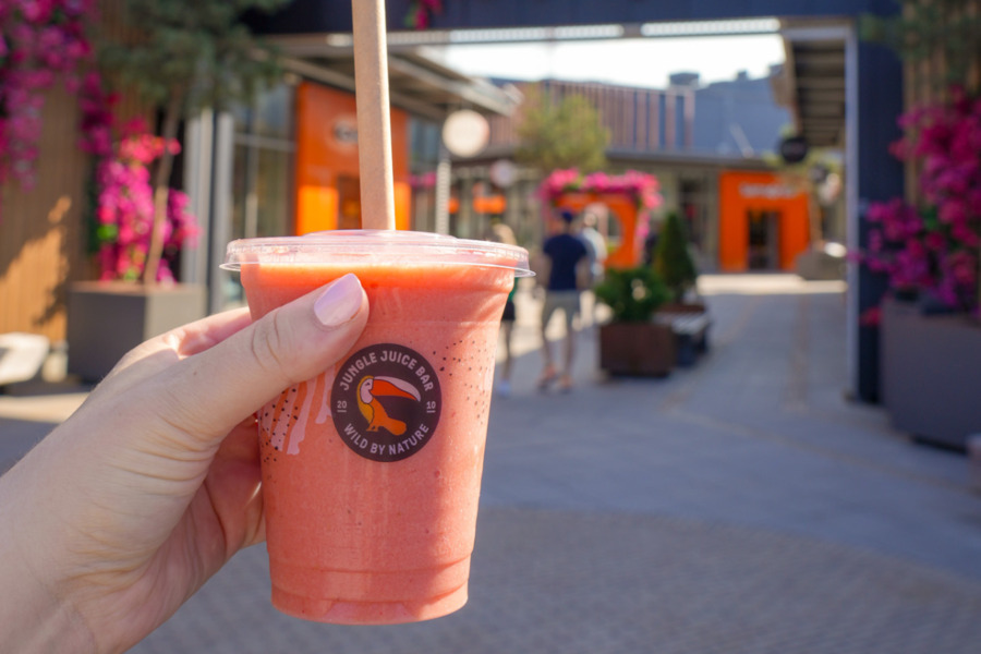 Jungle Juice Bar A hand holds a Jungle Juice Bar smoothie at the entrance of Helsinki Outlet Village on a sunny day.