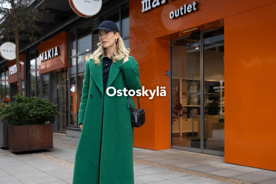 Vihreään takkiin ja mustaan lippikseen pukeutunut nainen vierailemassa ostoskylässä Marimekon ja Makian liikkeiden edessä