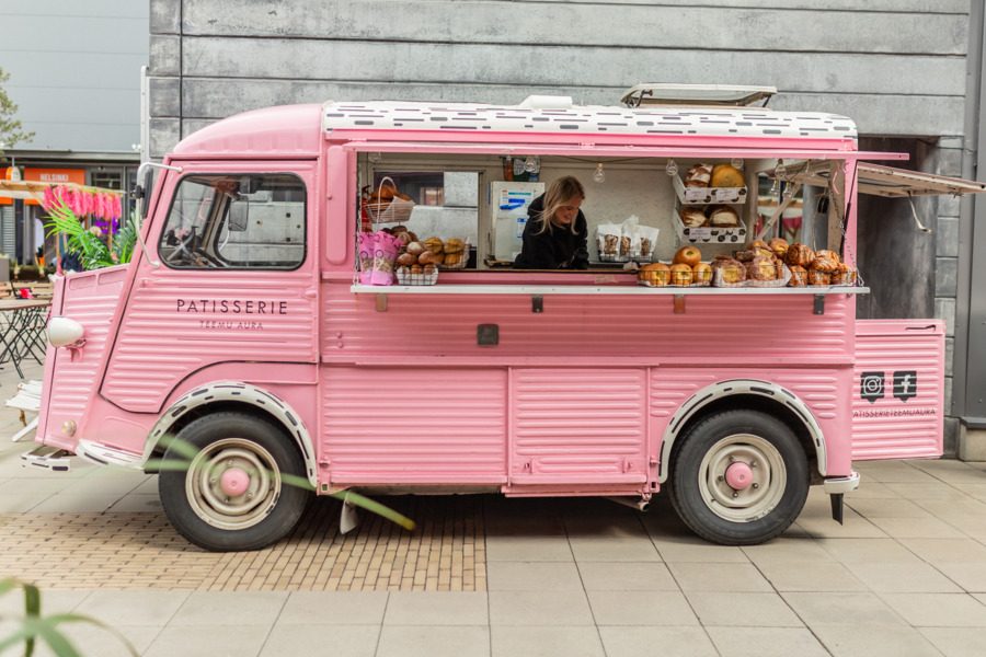 Pullabiili A pink bakery truck labeled "Patisserie" is parked on a paved area. The serving window is open, displaying various pastries, and a vendor is working inside.