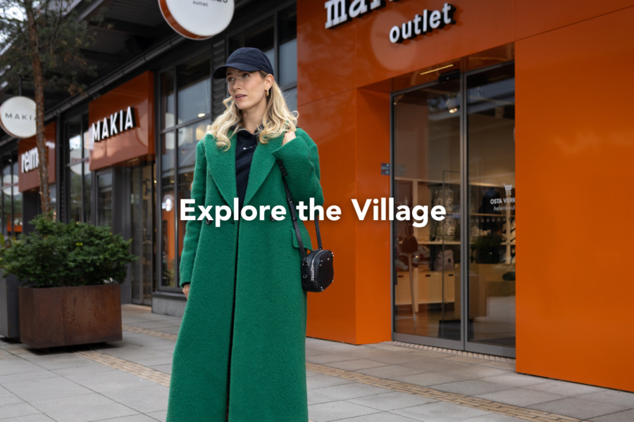 A woman wearing a green jacket and a black cap is shopping in the village in front of the Marimekko and Makia stores