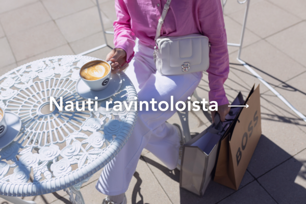 Nainen pinkissä paidassa ja valkoisissa farkuissa nauttii kahvia istuen valkoisessa puutarhakalusteella, käsissä ostoskasseja