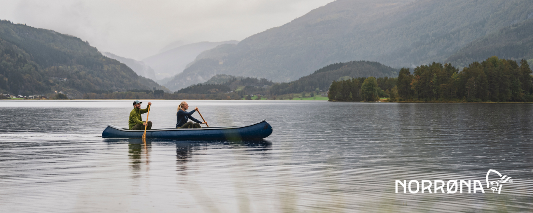A man and a woman are paddling a canoe in a mountainous landscape surrounded by green slopes
